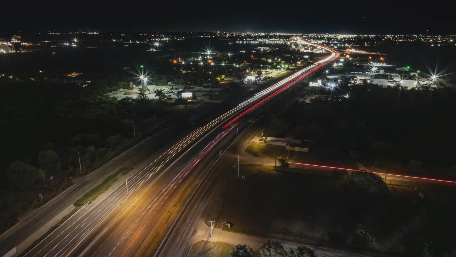 A long-exposure aerial view of a highway at night, showing streaks of red and white lights from vehicles with a brightly lit cityscape in the background.