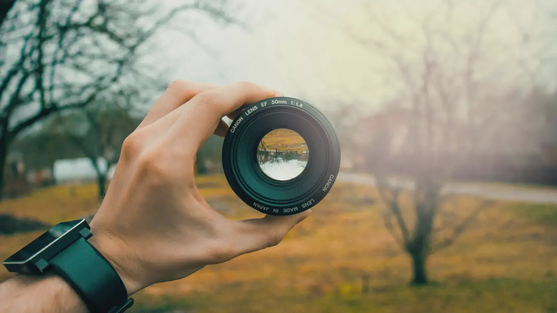 Hand holding a camera lens with sharp focus inside the lens and a blurred background, illustrating the concept of aperture and depth of field in photography.